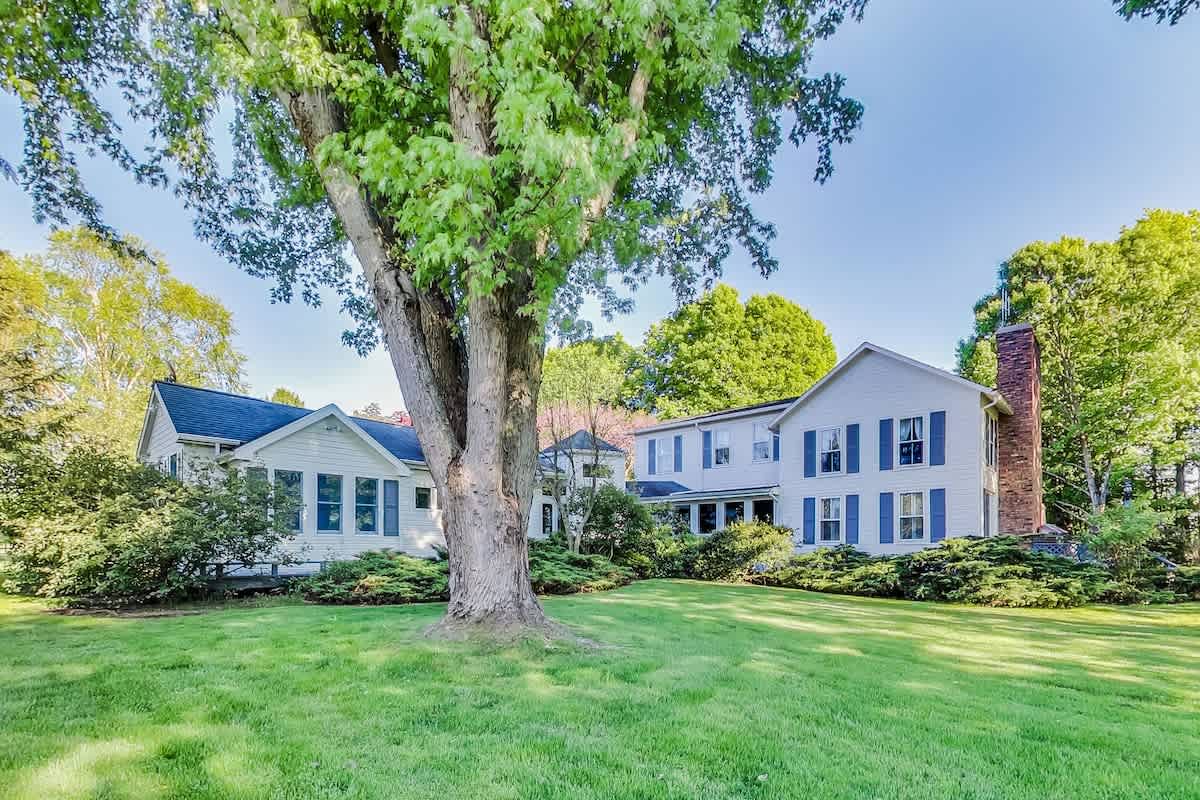 The back exterior of a large white farmhouse with blue shutters and a brick chimney, surrounded by a sprawling green lawn and mature trees, under a bright blue sky.