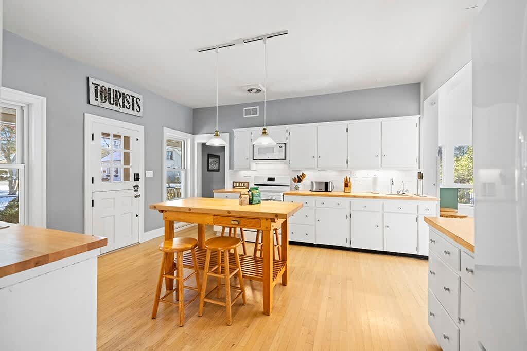 Bright kitchen with light gray walls, white shaker-style cabinets, light wood countertops, and a central wooden island with stools, featuring a white oven and microwave, and hardwood floors.