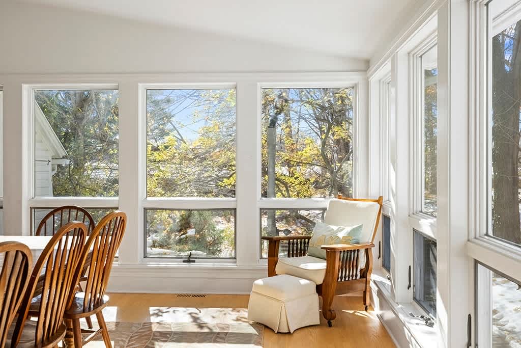 Bright dining area or sunroom with large windows offering views of the snowy outdoors, featuring light hardwood floors and a wooden dining table with matching chairs, alongside a comfortable armchair.