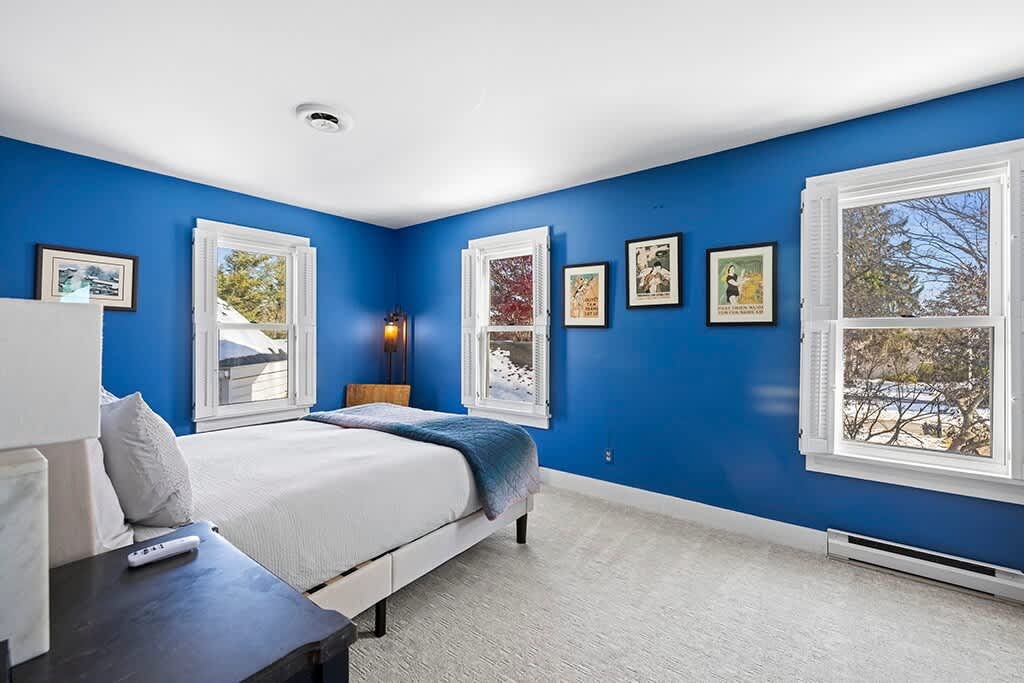 Another perspective of the blue-walled bedroom, showing the bed, a nightstand with a lamp, and more framed artwork. The room has a bright and airy feel with natural light from the windows.