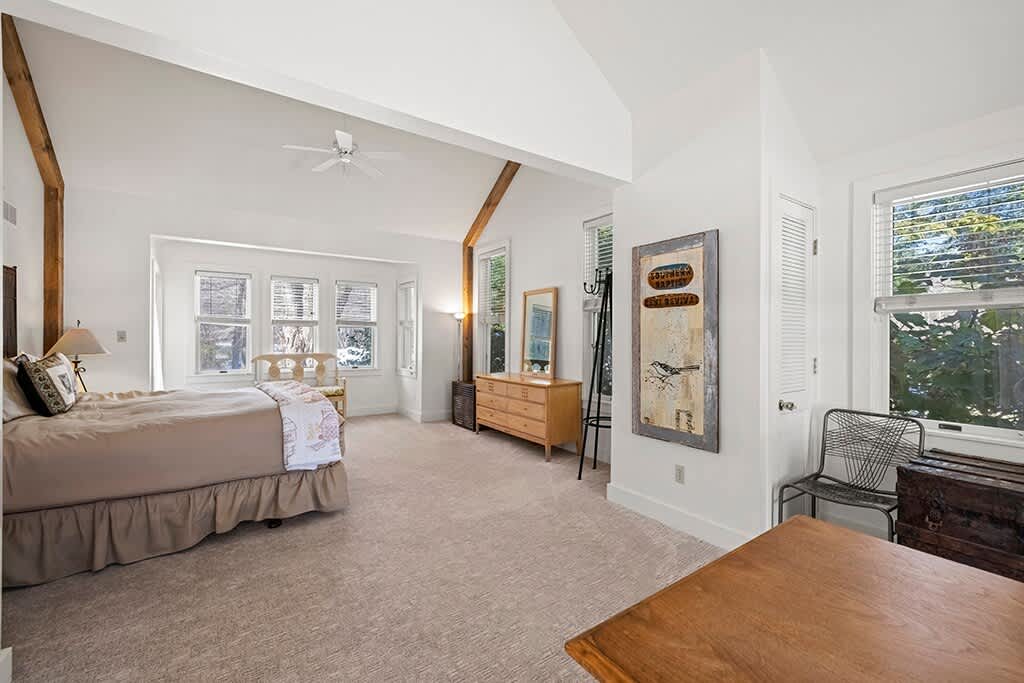 Wide view of a bright bedroom with light carpet, a king-sized bed, and a vaulted ceiling with exposed beams, showcasing multiple windows and a wooden dresser.