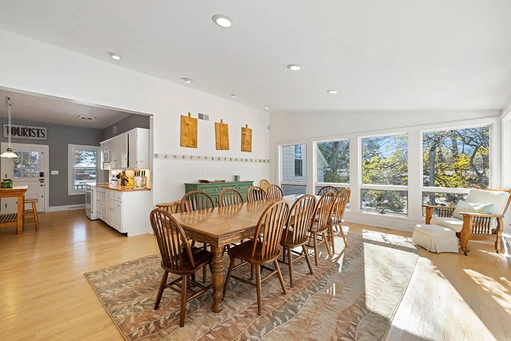 Expansive dining area with a long wooden table and multiple spindle-back chairs on a patterned rug, flowing into a white kitchen with a green island and a 'TOURISTS' sign above the doorway.