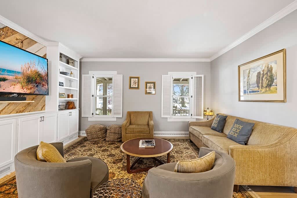 A comfortable living room featuring a gold sofa with throw pillows, two gray swivel chairs, and a round wooden coffee table on a patterned area rug. Built-in white shelving surrounds a TV mounted on a rustic wood accent wall, with windows featuring white shutters.