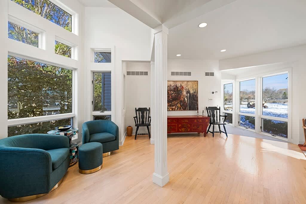 Bright living area with two teal swivel armchairs and a matching ottoman, positioned by expansive floor-to-ceiling windows that offer abundant natural light and views of the outdoors. A red console table with abstract art hangs on the wall.