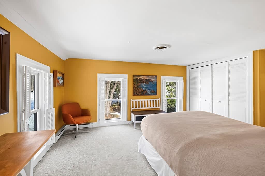 Wide view of the yellow-walled bedroom showing a bed, an orange accent chair, a long white bench, and a wall of white louvered closet doors.