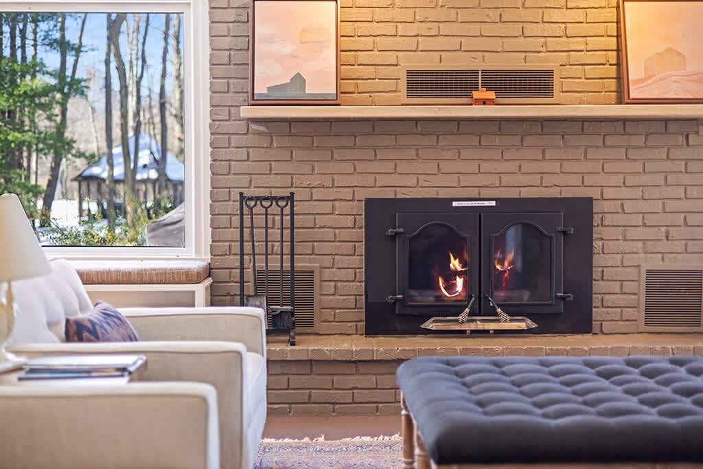 Close-up view of the brick fireplace with a black insert and a wooden mantel, flanked by large windows offering a view of trees, with a black fireplace toolset beside it.