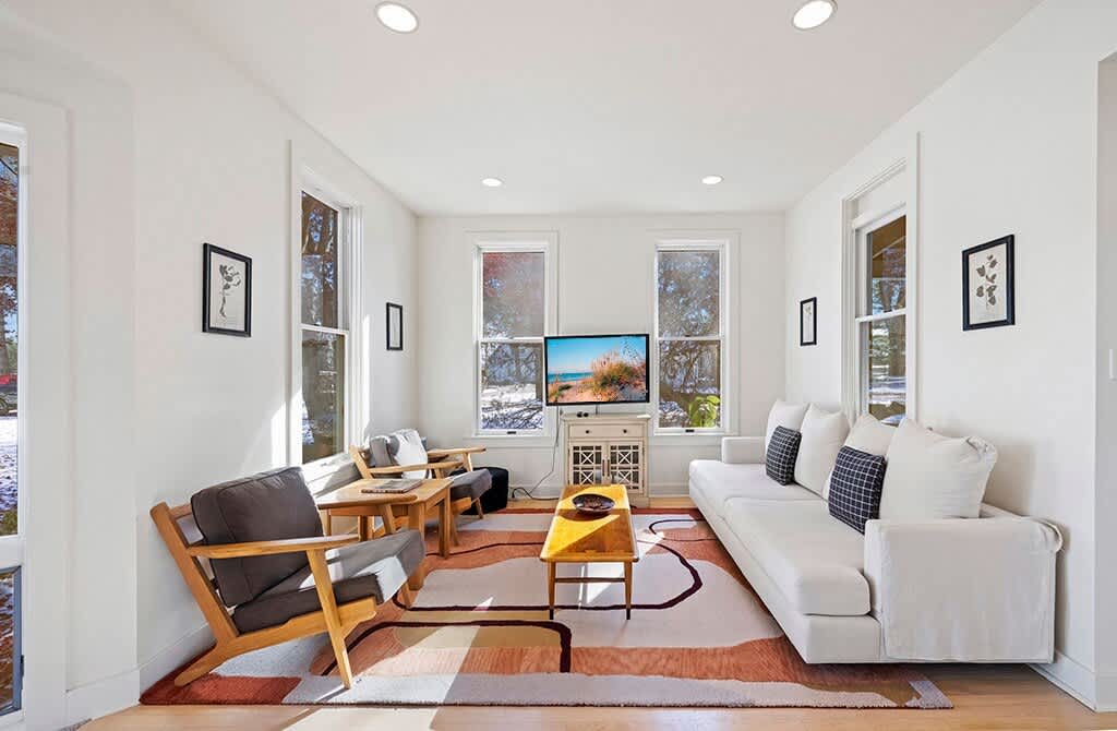 A bright living room with light hardwood floors and white walls, featuring a white sofa with throw pillows, two wooden armchairs with dark cushions, and a wooden coffee table on a geometric area rug. Large windows provide natural light and views of trees.
