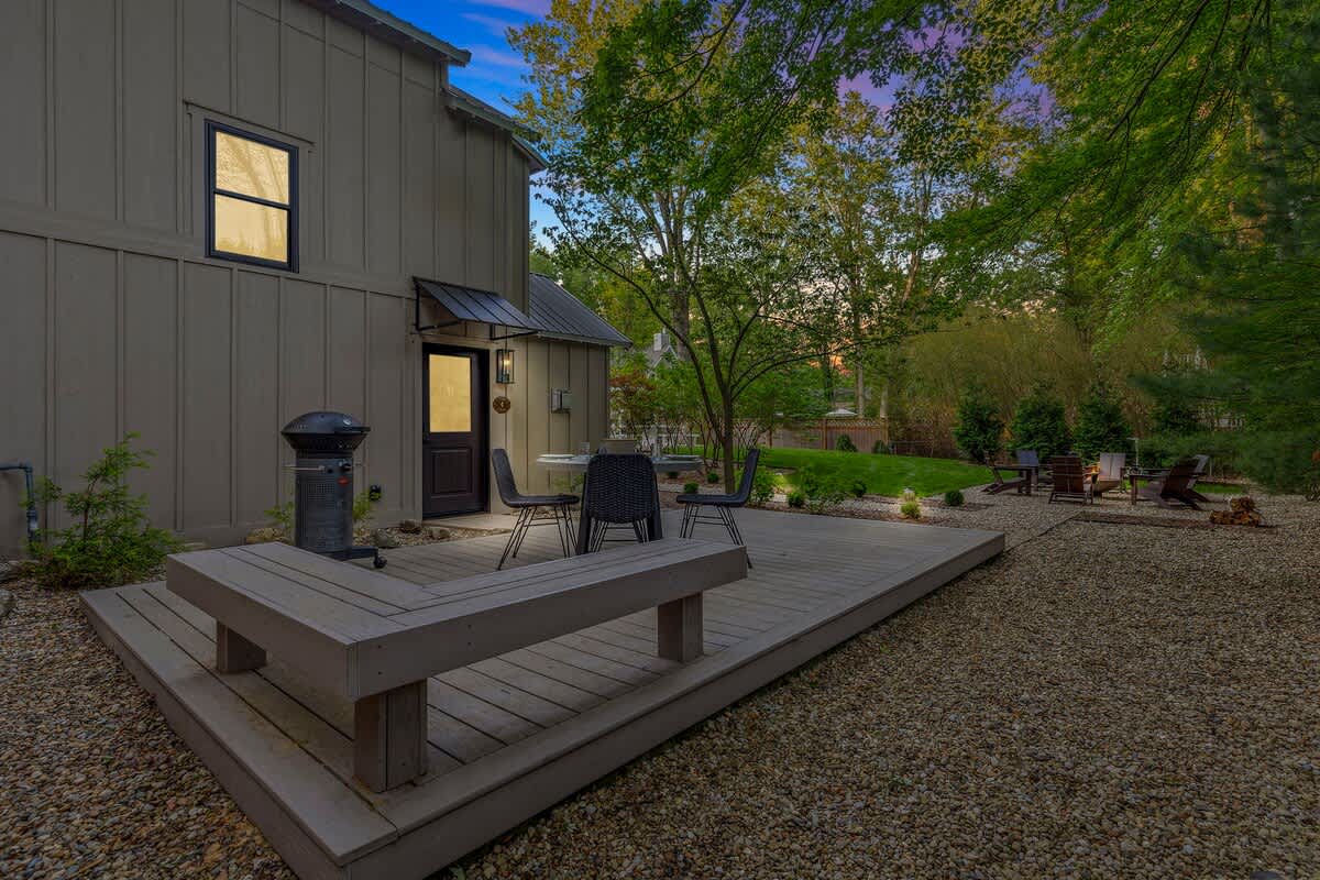 An evening view of the house's back exterior, highlighting the wooden deck with an integrated bench, an outdoor dining set, and a BBQ grill, all situated on a gravel yard with a distant fire pit area and lush trees under a twilight sky.