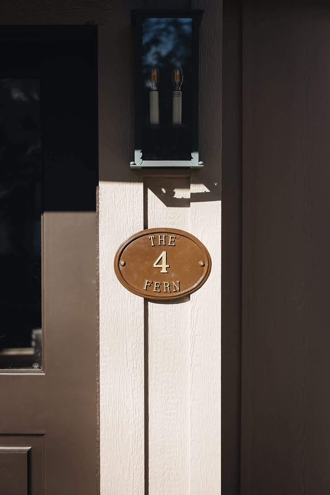 Close-up of a bronze oval sign on a light-colored wall, displaying "THE 4 FERN" in raised lettering, next to a dark exterior light fixture.