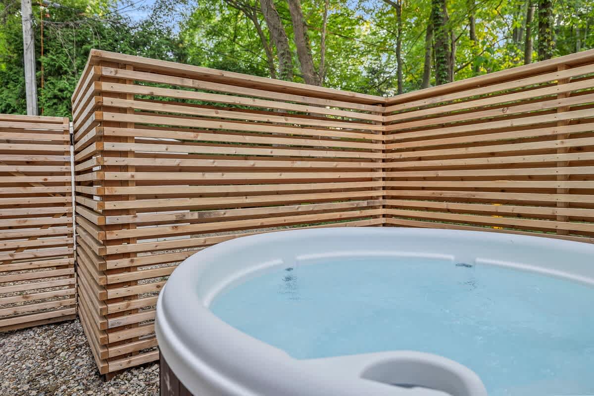 A white hot tub filled with sparkling water is positioned on a gravel surface, enclosed by a stylish slatted wooden privacy fence, with green trees visible in the background.