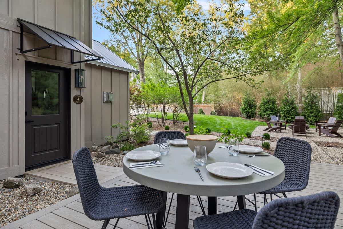 A close-up of the outdoor dining area on the wooden deck, featuring a round concrete-style table with four woven chairs, set with plates and glasses, adjacent to the house's dark entrance door with a small awning.