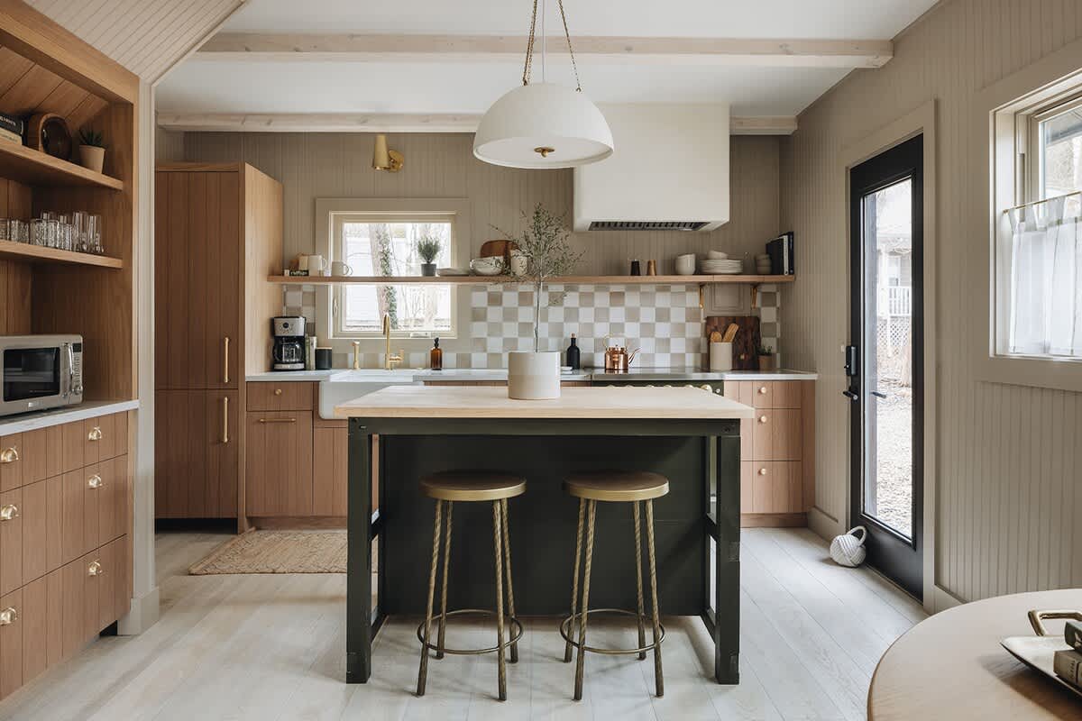 Bright kitchen with light wood cabinets, a dark green island with brass bar stools, a white farmhouse sink, and a checkered tile backsplash. Light wood floors and a pendant light complete the space.