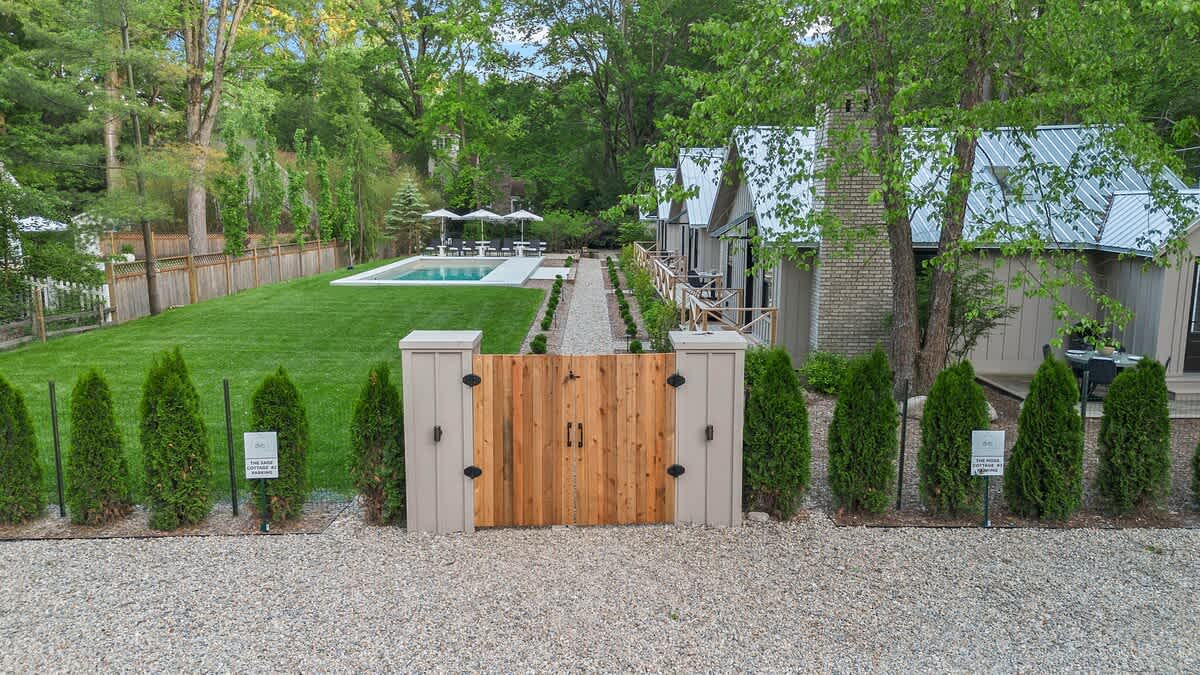 A ground-level view of the vacation rental property entrance, featuring a wooden gate flanked by small evergreen shrubs, leading to a gravel path and several light-colored cottages with metal roofs.
