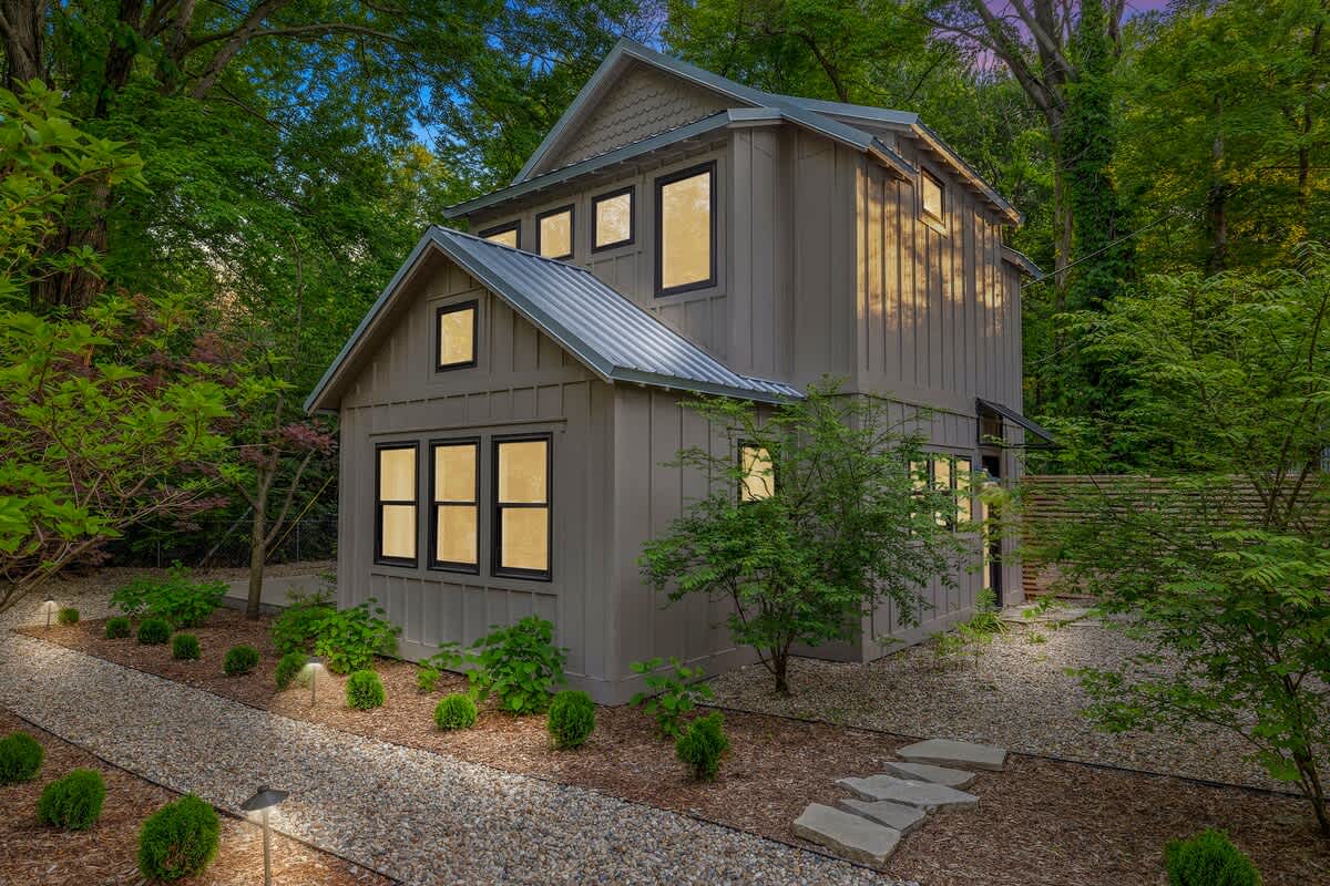 The front exterior of a tall, modern board and batten house with a metal roof and numerous black-framed windows, illuminated by warm interior lights at dusk, with a gravel path and landscaped shrubs leading to the entrance.