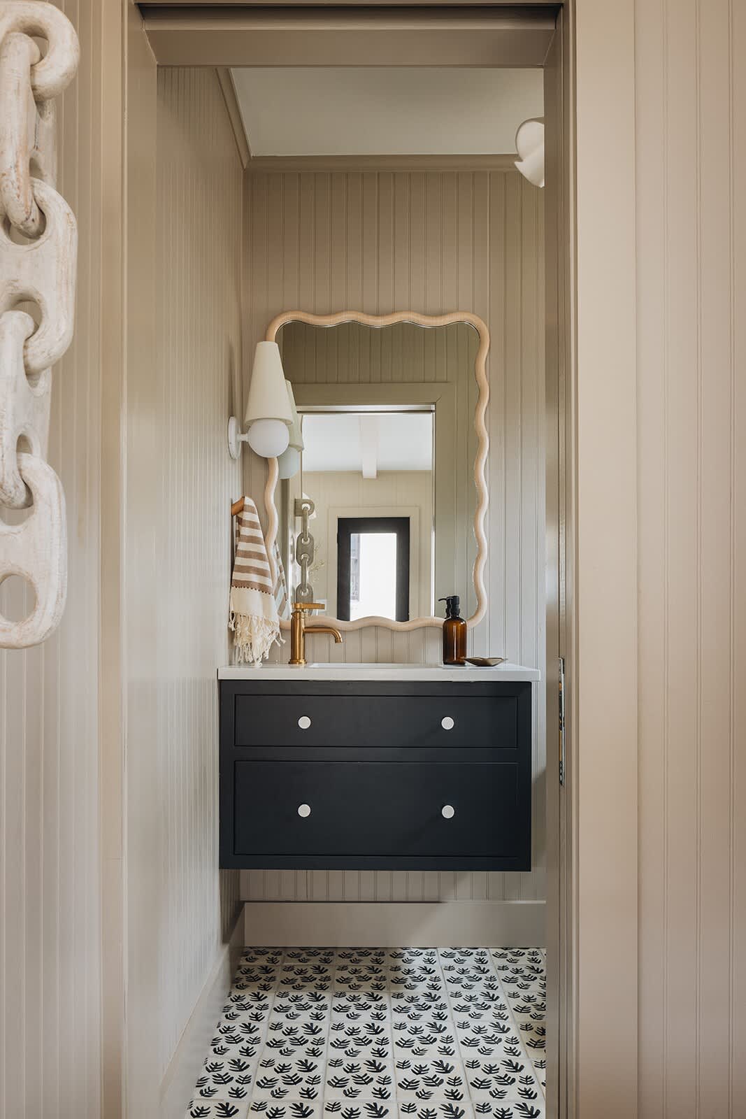 A stylish powder room with a black floating vanity, white knobs, a white countertop, a wavy-framed mirror, and a wall sconce, with patterned floor tiles and beadboard walls.