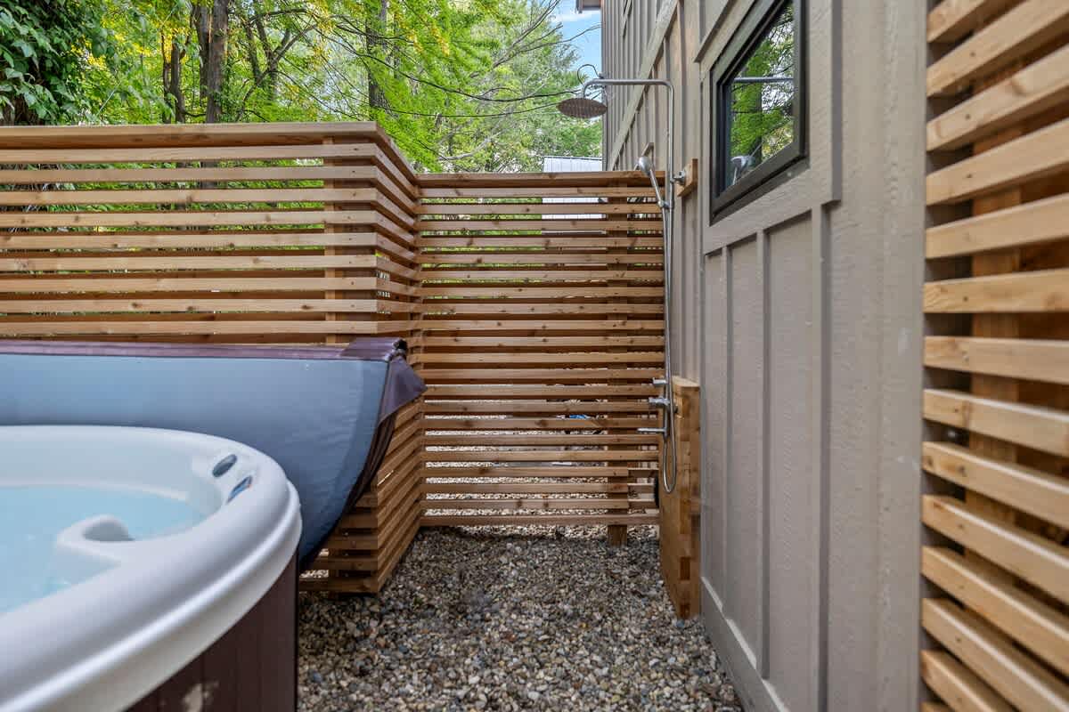 The hot tub area features a slatted wooden privacy fence and an outdoor shower mounted on the house's exterior, all set on a gravel ground, providing a convenient rinse-off spot.