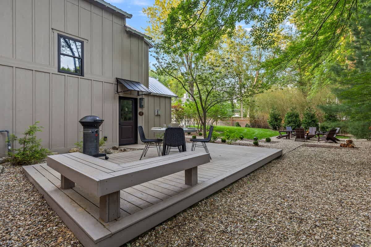 A wide view of the house's back exterior, showcasing a large wooden deck with an integrated bench, an outdoor dining set, and a BBQ grill, all set on a gravel yard with a grassy area and mature trees in the background.