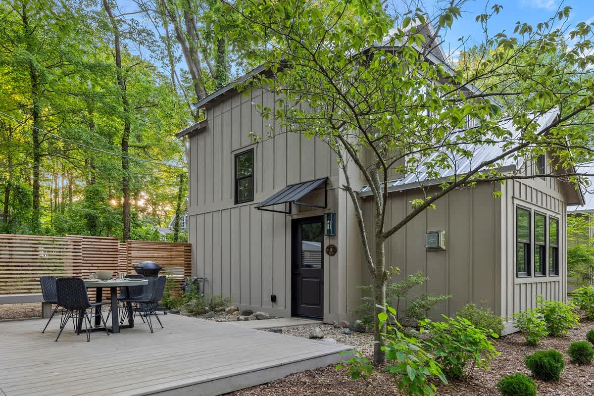 The back exterior of a modern board and batten house with a metal roof, featuring a spacious wooden deck with an outdoor dining set and a BBQ grill, surrounded by lush trees and gravel landscaping.