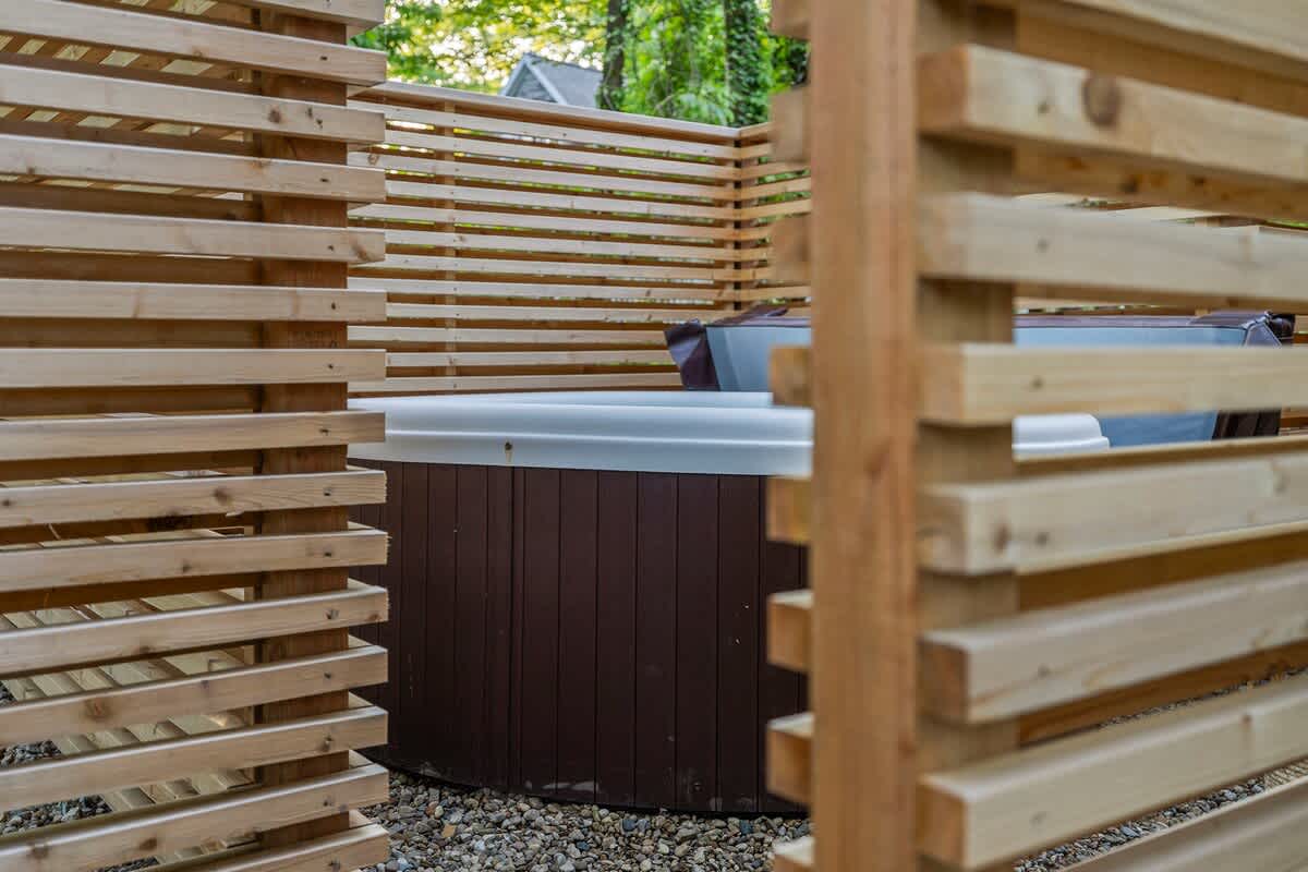 A hot tub is partially visible behind a modern slatted wooden privacy fence, set on a gravel ground, suggesting a secluded outdoor relaxation area.