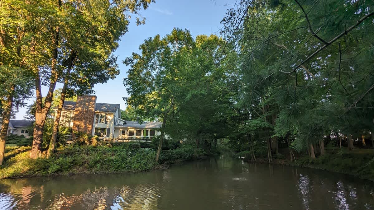 A large, multi-story house with a prominent brick chimney and numerous windows stands beside a tranquil body of water, framed by a dense line of tall, mature trees under a clear sky.