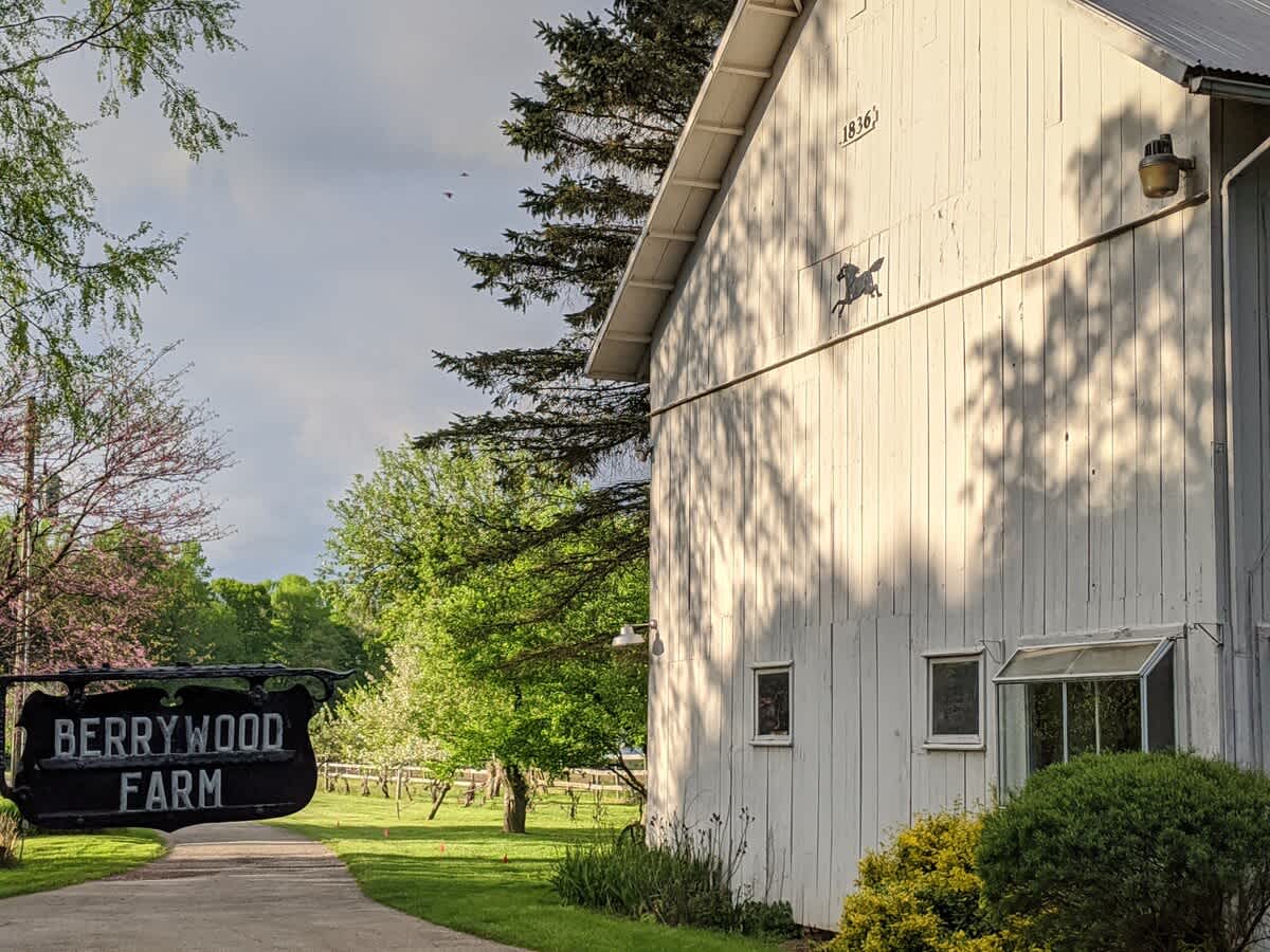 A white barn-like building with "1836" and a horse silhouette on its side stands beside a paved path, with a black "BERRYWOOD FARM" sign in the foreground and lush trees in the background.