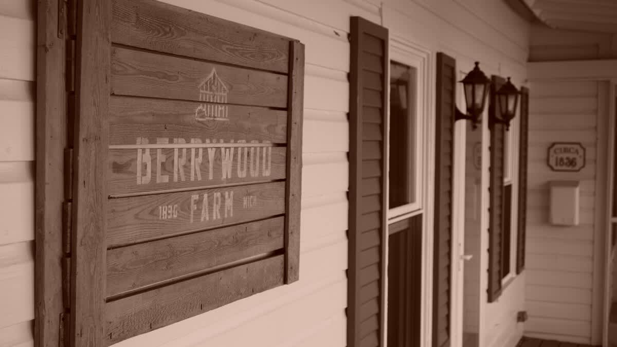 A sepia-toned image shows a rustic wooden sign with "BERRYWOOD FARM" and "1836" mounted on white horizontal siding, flanked by dark window shutters and traditional outdoor lanterns.