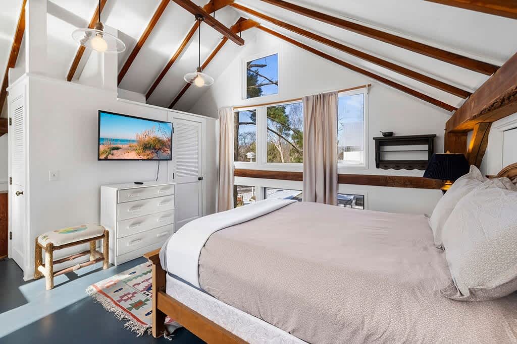Bedroom area with a queen bed, a white dresser with a mounted TV, and a small stool. The room has a vaulted ceiling with exposed beams and large windows, with a colorful rug on the dark floor.