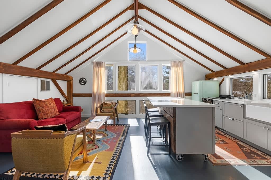 Bright, open-concept loft space with a red sofa and armchairs on a vibrant rug, facing a modern kitchen island with bar stools. The room features a high vaulted ceiling with exposed wooden beams and large windows providing ample natural light.