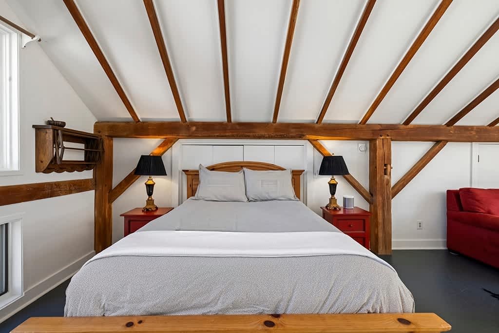 Cozy bedroom area within the loft, featuring a queen-sized bed with light gray bedding, flanked by red nightstands with black lamps. Exposed wooden beams frame the bed, and a vaulted ceiling adds to the spacious feel.