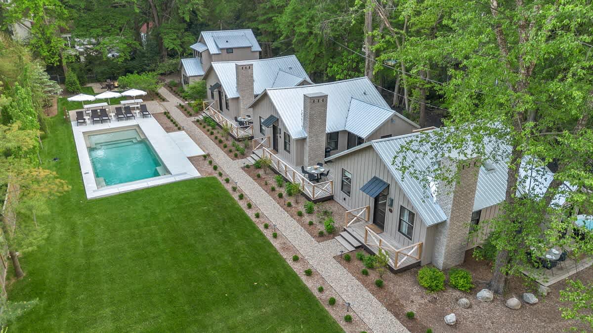 Overhead aerial view showcasing multiple modern cottages with light siding and metal roofs, connected by gravel paths, alongside a large swimming pool and manicured lawn surrounded by lush trees.