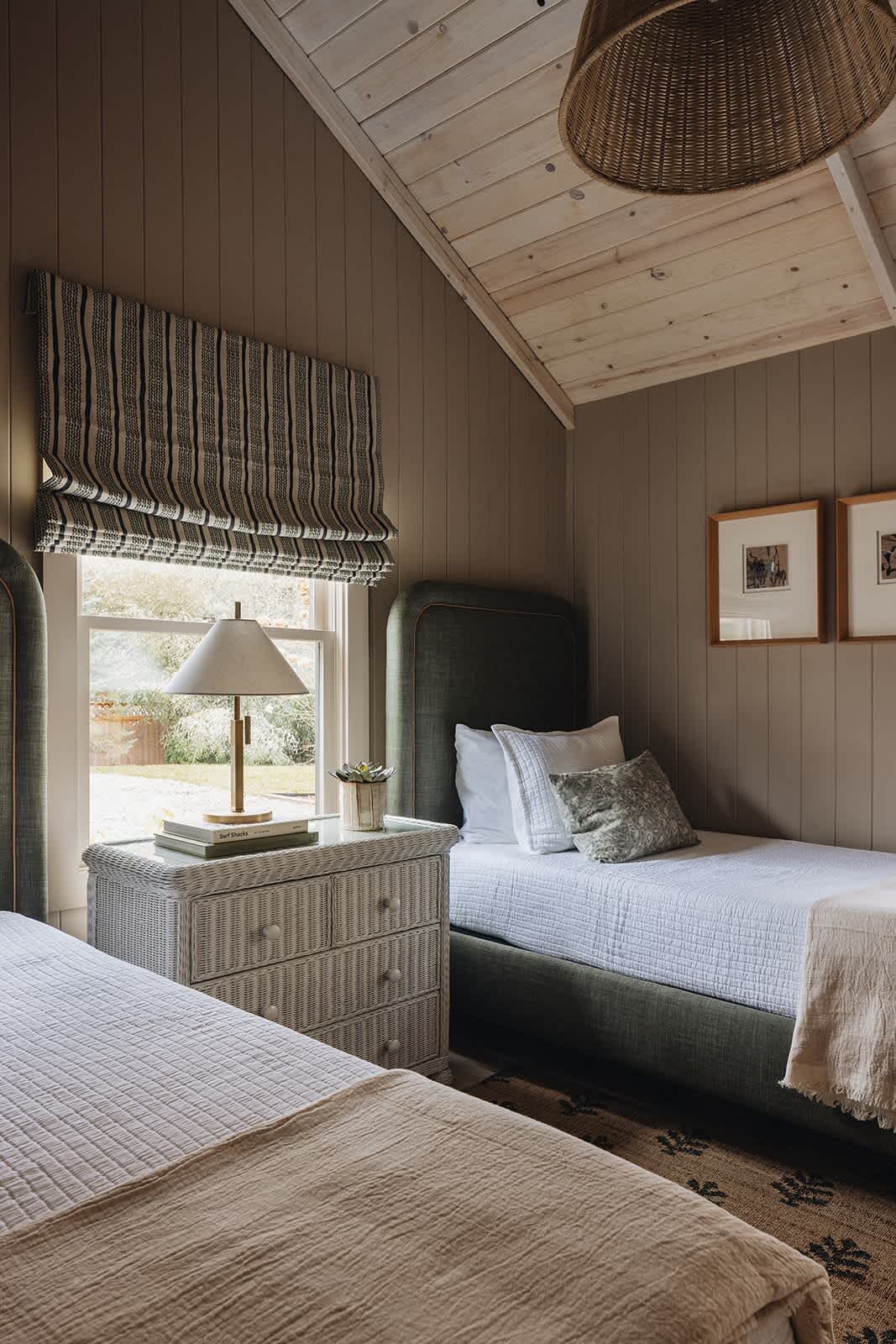 View of a twin bedroom with green upholstered beds, a white wicker nightstand with a lamp, and light brown vertical plank walls, featuring a vaulted wooden ceiling and framed artwork.