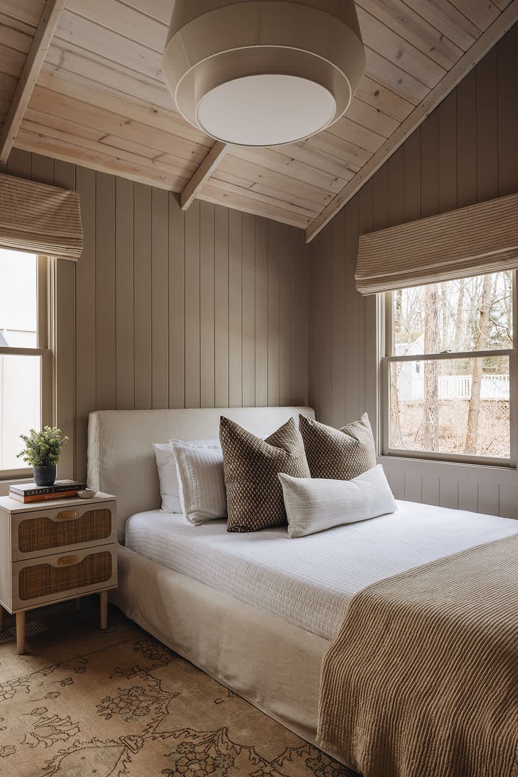 Cozy bedroom featuring a single bed with a cream upholstered headboard, a woven nightstand, and light brown vertical plank walls, complemented by a vaulted wooden ceiling and a large drum pendant light.