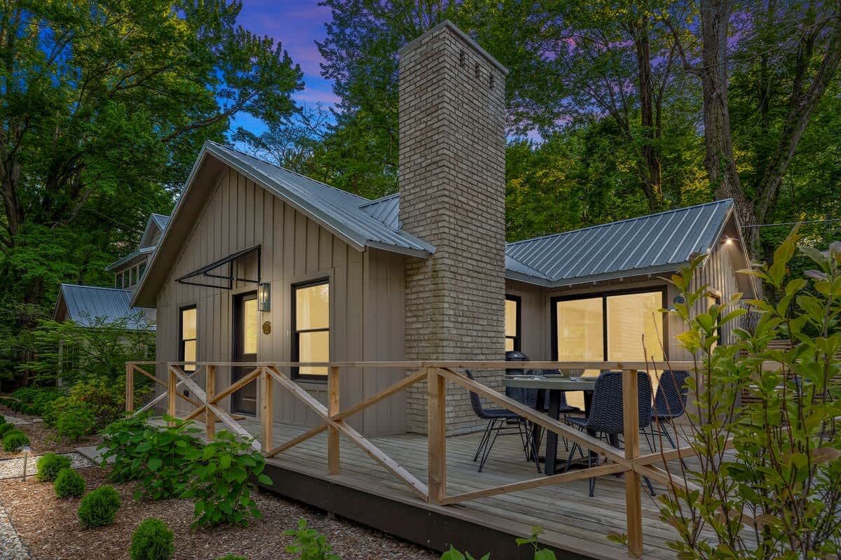 Modern cottage with light brown vertical siding, a grey metal roof, and a prominent brick chimney, featuring a wooden deck with a dining set and surrounded by lush green trees.