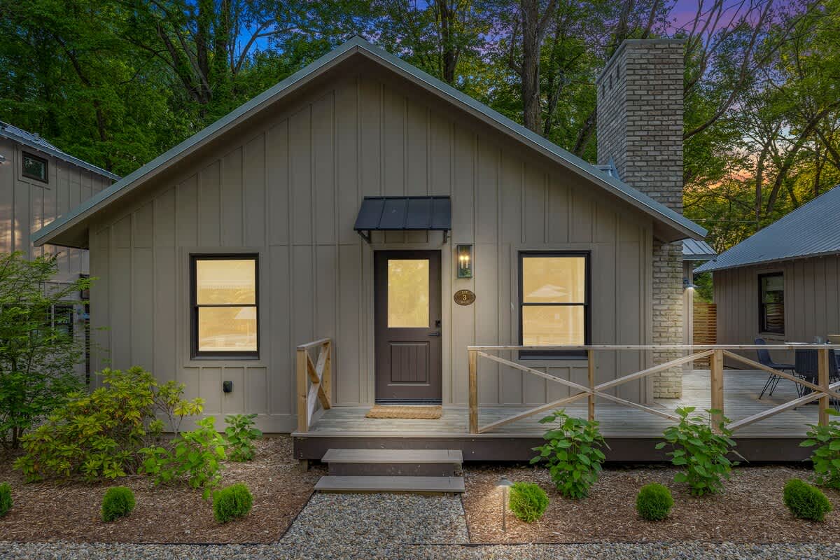 Front exterior of a charming cottage with light brown vertical siding, a dark brown front door with a small awning, and a wooden porch with railings, surrounded by landscaping.