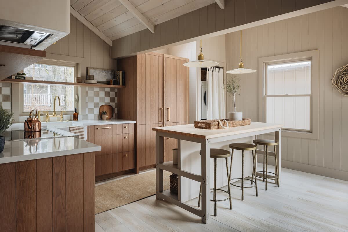 A bright kitchen with light wood cabinetry, a checkered tile backsplash, and a large island with bar stools, featuring vaulted wood ceilings and light wood flooring. A laundry area is visible in the background.