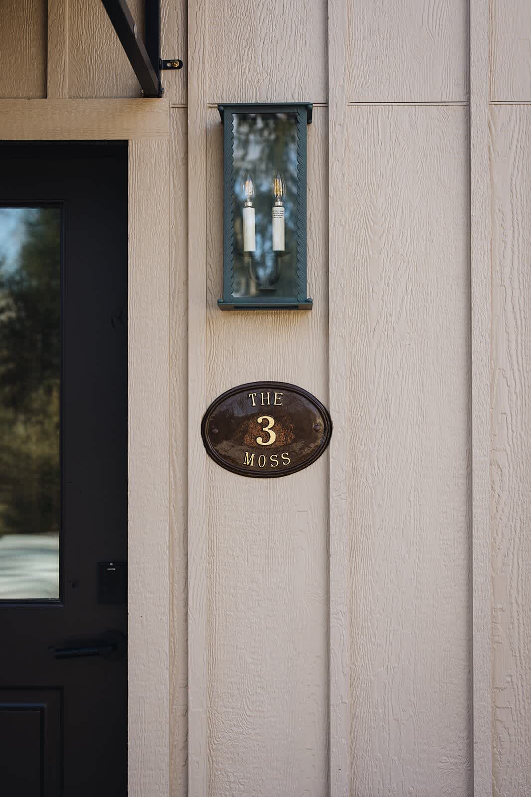 Close-up of a dark brown oval plaque with 'THE 3 MOSS' in gold lettering, mounted on light beige board-and-batten siding next to a black door and a wall-mounted lantern.