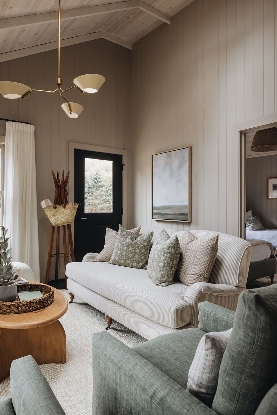 A bright living room featuring a striped sofa with decorative pillows, a wooden coffee table, and a modern chandelier, all under a high vaulted wood ceiling with light beige vertical paneling walls.