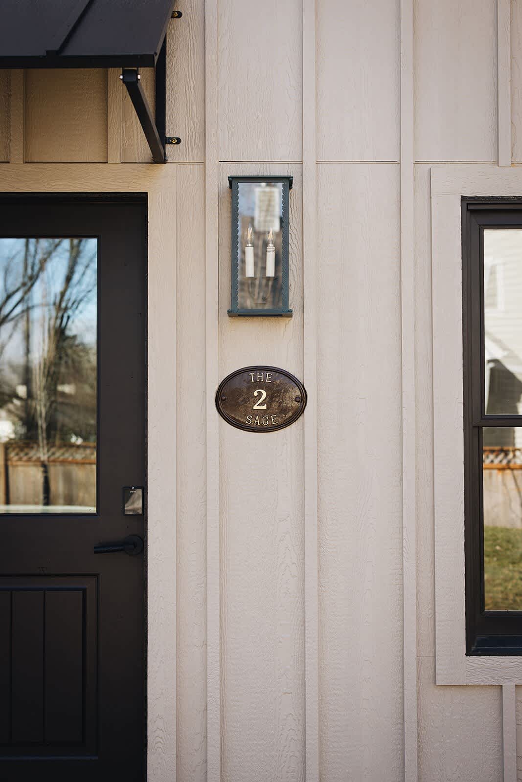 A close-up of the cottage's front entrance, showing a dark brown door with a black awning and a vintage-style oval plaque displaying "2 SAGE". The light beige board-and-batten siding and an outdoor wall lantern are also visible.