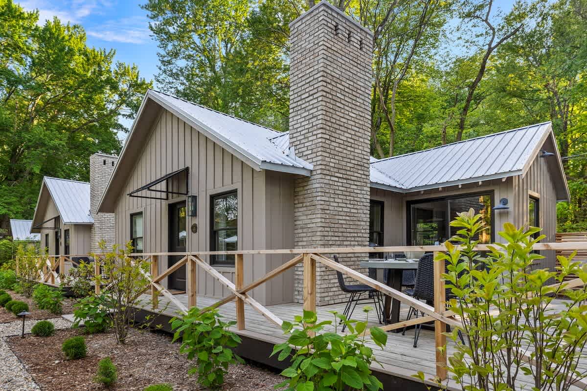 A side view of a modern cabin with light beige siding and a metal roof, featuring a prominent brick chimney. A wooden deck extends along the side, furnished with a small outdoor dining table and chairs, and surrounded by wooden railings and young plants.