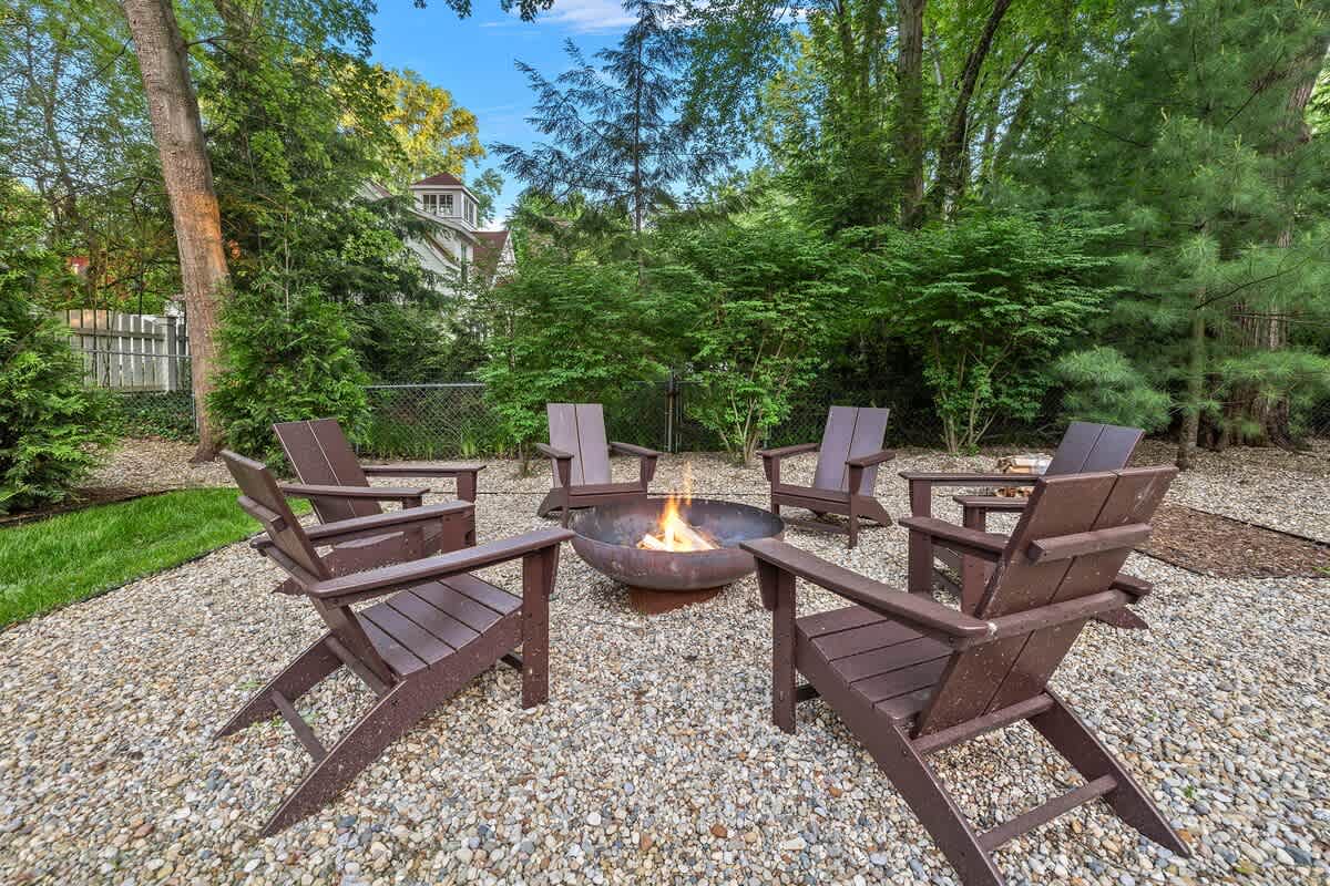 A circular fire pit with a burning fire is surrounded by six dark brown Adirondack chairs on a gravel patio, nestled within a lush green yard with mature trees.
