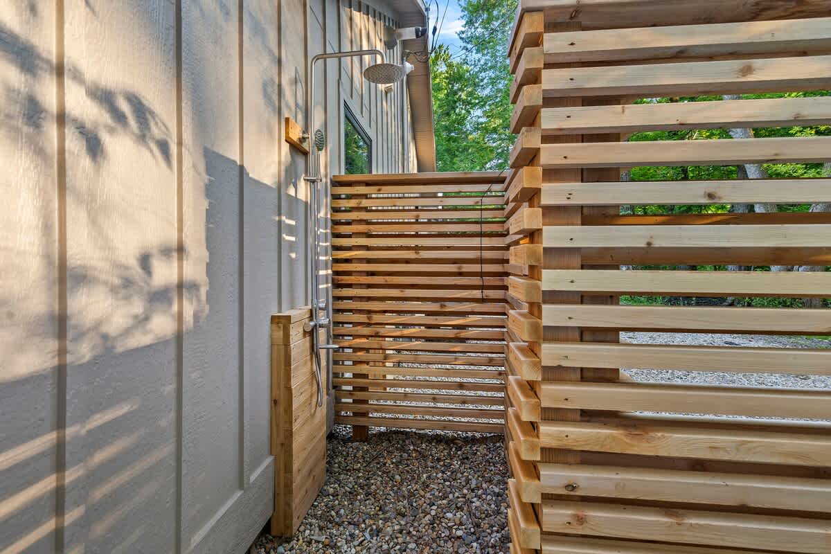 An outdoor shower with a chrome shower head is mounted on a light-colored wooden wall, enclosed by a wooden slatted privacy screen and set on a gravel ground, with trees in the background.