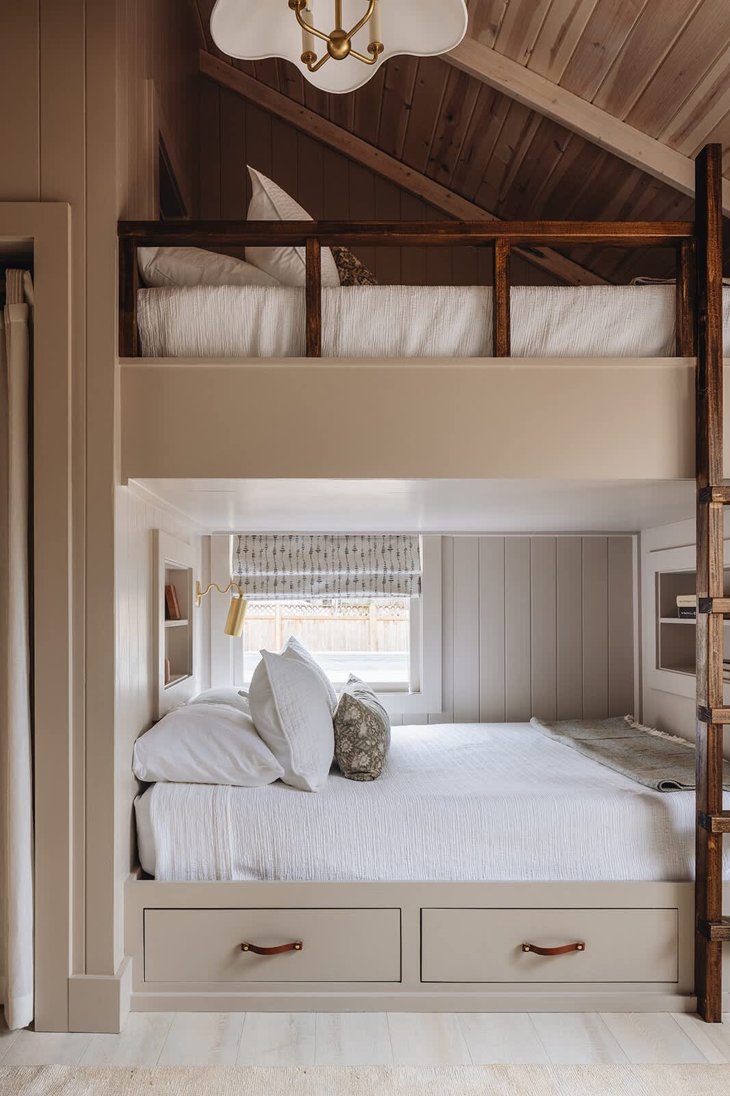 A cozy bedroom featuring a built-in bunk bed structure with a queen-sized bed on the bottom and a twin bed on top. The room has light-colored vertical wood paneling, a vaulted ceiling, and built-in storage drawers.