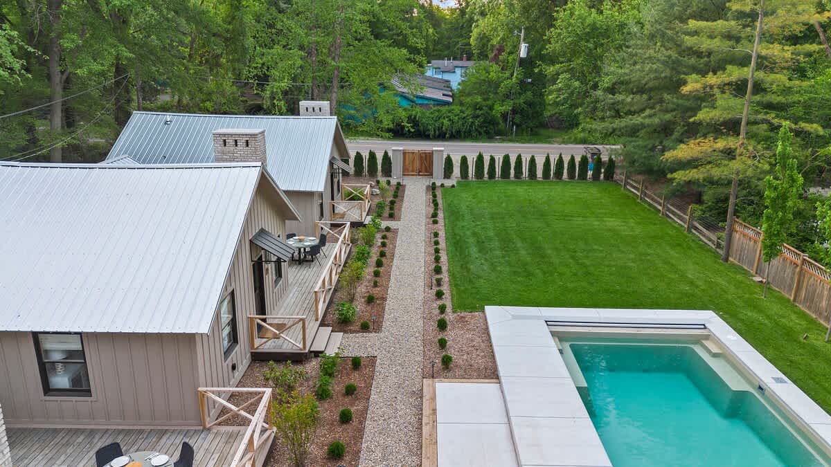 An aerial shot looking down the length of the property, showing a series of modern cabins with light siding and metal roofs, connected by a gravel path. A large green lawn stretches to the right, ending at a rectangular swimming pool with a white surround.