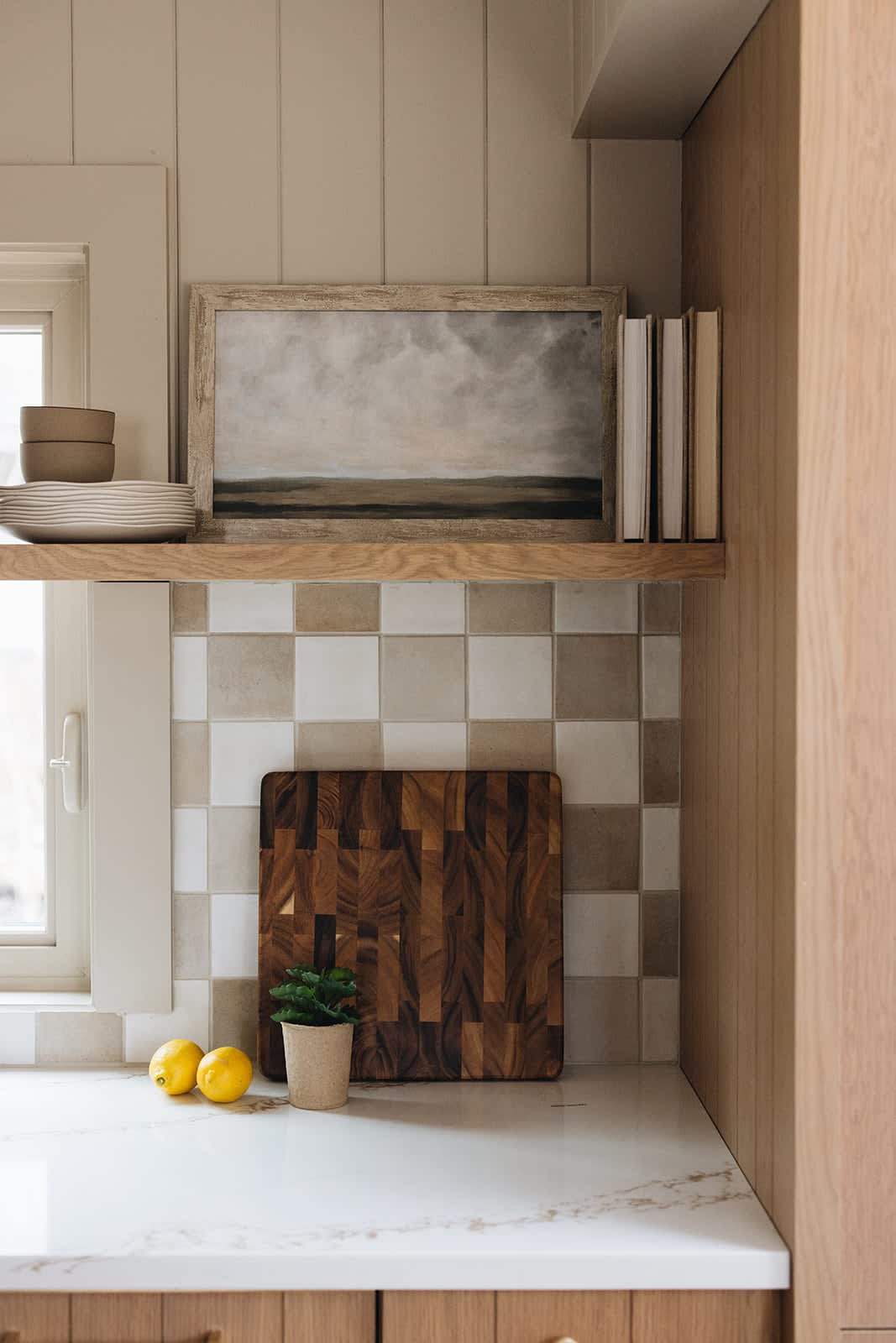 Close-up of the kitchen counter with a checkered tile backsplash, a wooden cutting board, and two lemons. A framed landscape painting and books are displayed on a floating wooden shelf above.