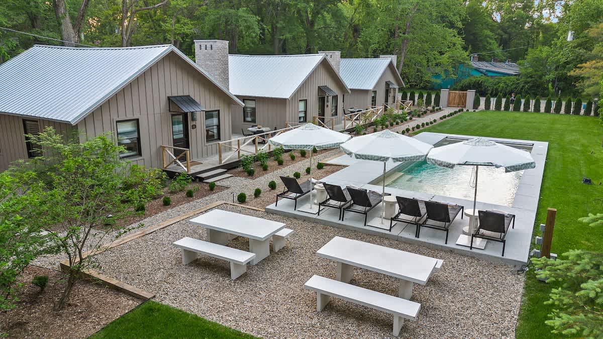 An elevated view of the property featuring a rectangular swimming pool with lounge chairs and umbrellas, alongside two white picnic tables on a gravel patio. Several modern cottages with light siding and metal roofs are visible in the background, surrounded by green lawn and trees.