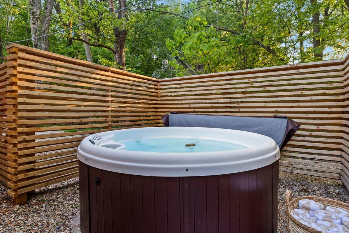 A bubbling hot tub is nestled within a private enclosure made of horizontal wooden slats, set on a gravel surface. Lush green trees provide a natural backdrop, enhancing the secluded and relaxing atmosphere.
