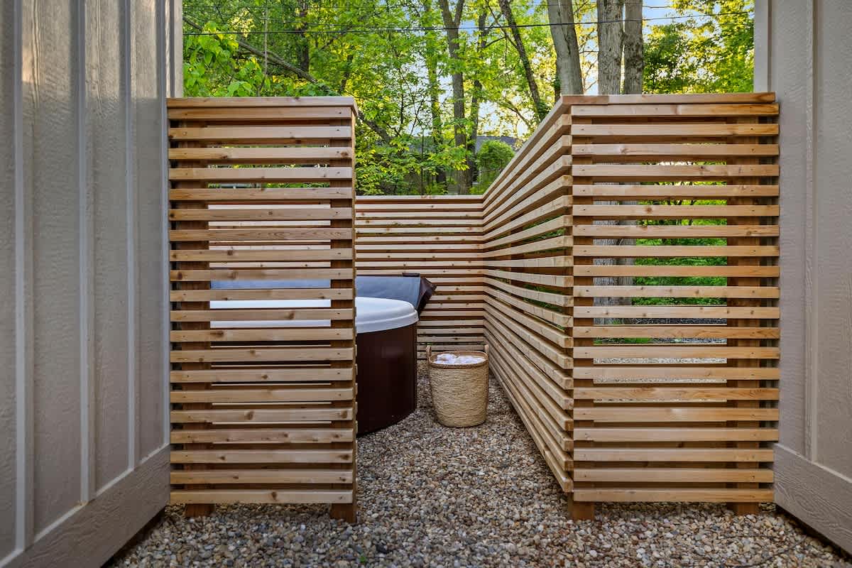 A private hot tub area is enclosed by modern slatted wooden screens, creating a secluded space on a gravel ground. A dark brown hot tub with a light cover is visible, along with a woven basket containing white towels.