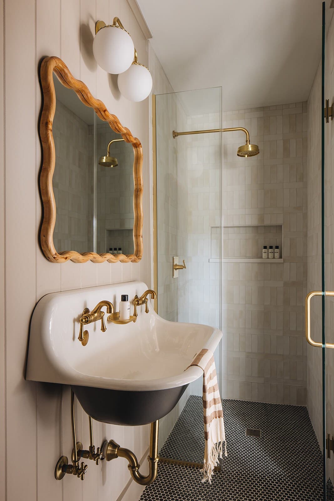 A modern bathroom with a walk-in shower featuring light rectangular tiles and brass fixtures. The wall-mounted double basin sink with brass fixtures and a wavy wooden mirror are also visible.