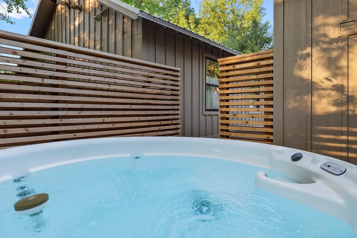 A white hot tub with light blue water is positioned outdoors, surrounded by modern horizontal wooden slatted privacy screens, with a rustic building and green trees in the background.