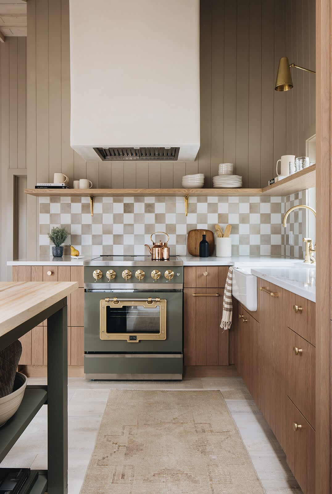 A stylish kitchen corner featuring a sage green range oven with brass accents, set against a checkered tile backsplash in neutral tones. Light wood cabinets with brass pulls, open shelving, a farmhouse sink, and white countertops complete the rustic-modern aesthetic.